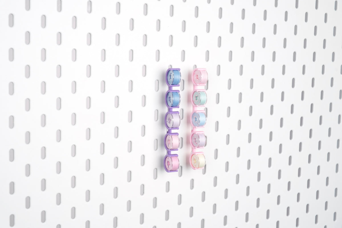 Two five-piece pegboard bobbin holder sets in violet purple and pink, storing ten sewing bobbins