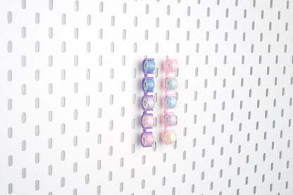 Two five-piece pegboard bobbin holder sets in violet purple and pink, storing ten sewing bobbins